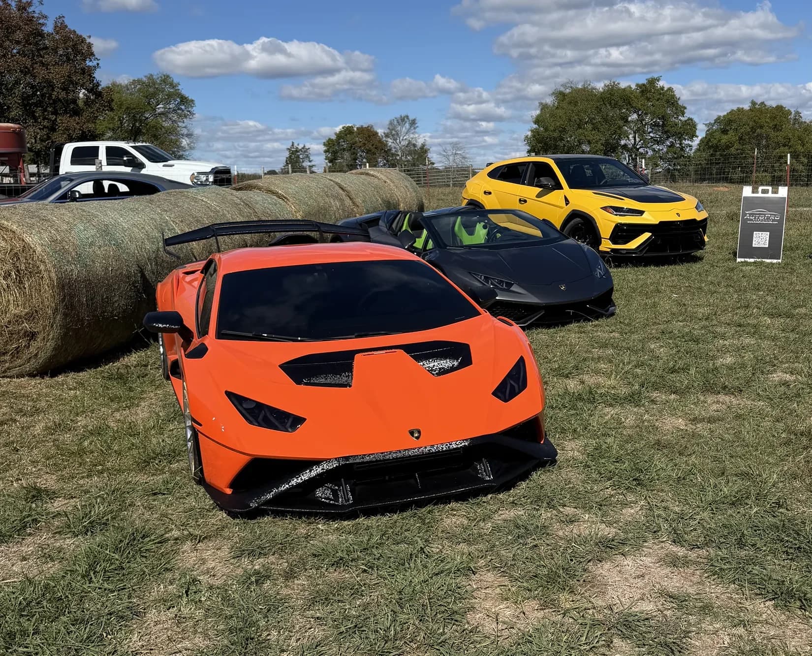 An orange, a black and a yellow Lamborghini on the grass beside hay bales on October 17