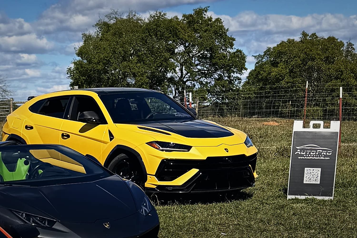 A yellow Lamborghini Urus on the grass at Rancho Jaramillo beside the AutoPro Nashville sign, a black Lamborghini in front of it
