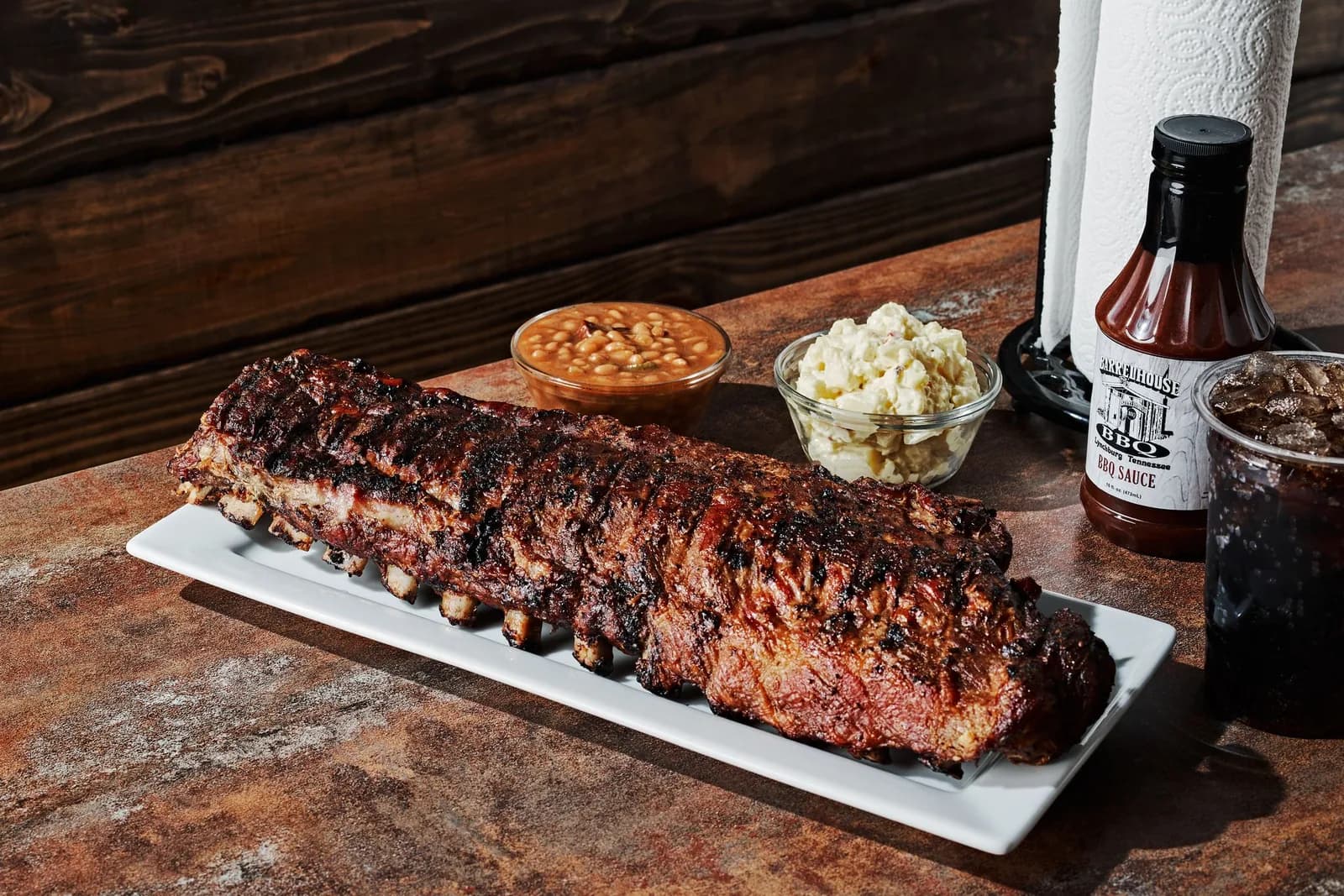 A full rack of ribs on a long platter beside baked beans, potato salad and a bottle of their sauce
