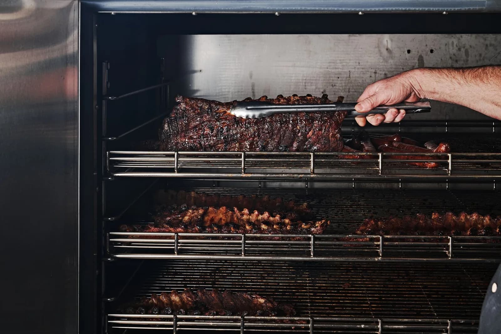 Ribs and sausage on the racks of a stainless steel smoker, a rack lifted with tongs