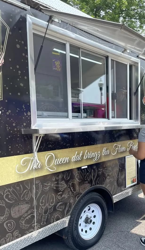 The left side of her black food trailer at an outdoor event, a gold band across it in white script reading Tha Queen dat bringz tha Flava, seafood line art below
