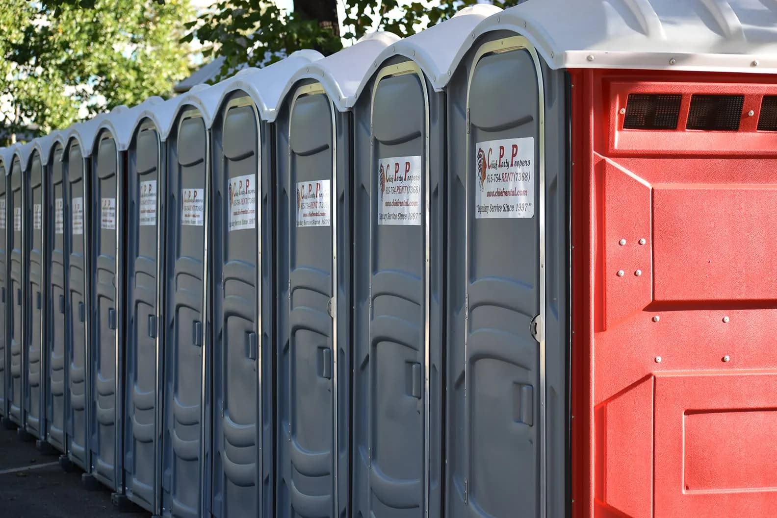 A long row of gray portable restrooms, the nearest with its red door to the camera