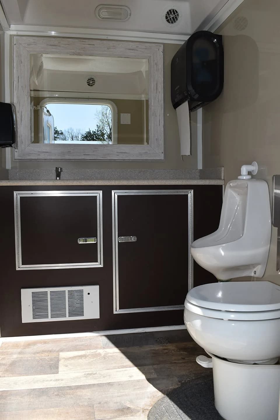 Inside a restroom trailer: a framed mirror over a vanity with dark cabinets, a urinal and a toilet