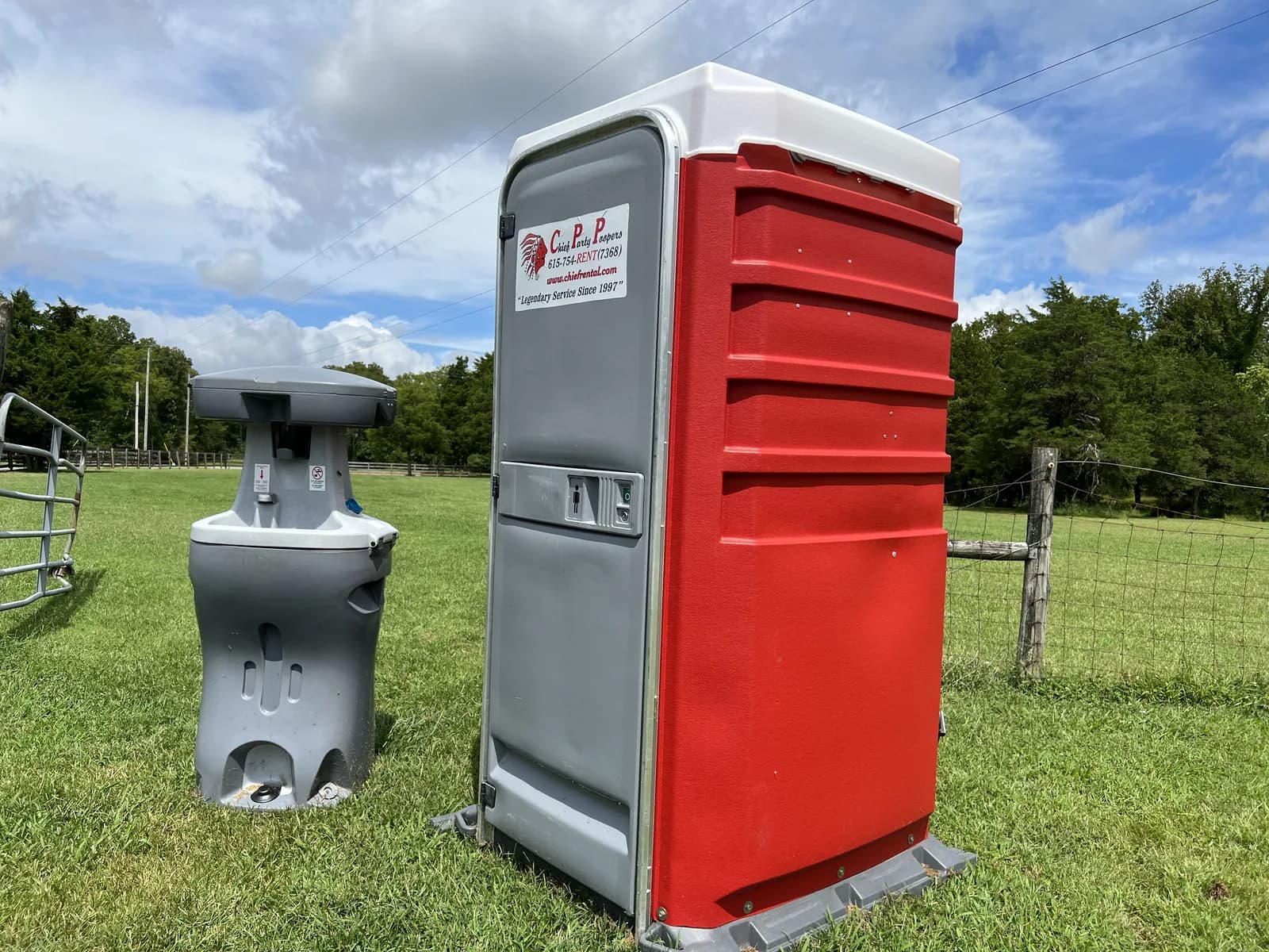 A red and gray portable restroom with a hand-washing station beside it, on the grass of a fenced pasture