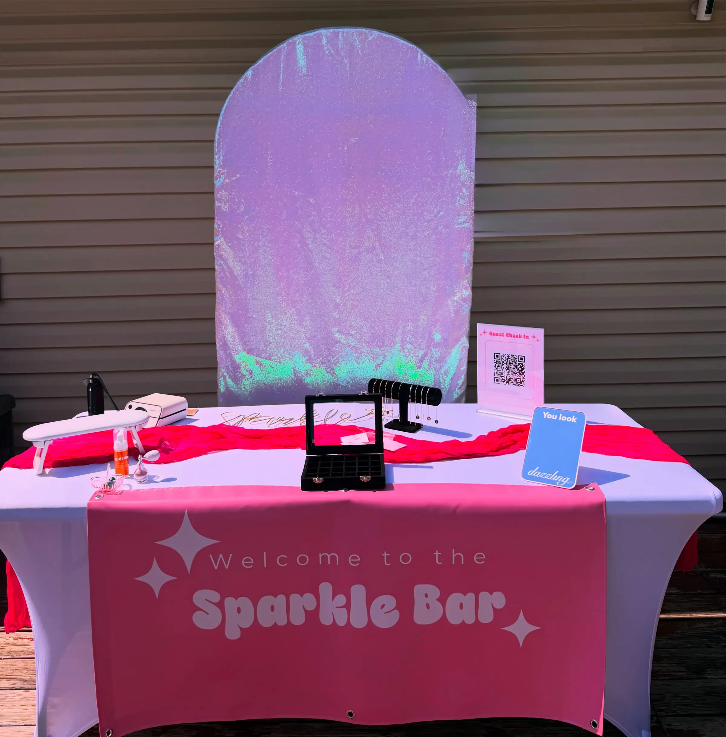 The Sparkle Bar booth set up outdoors: a Welcome to the Sparkle Bar table banner, an iridescent arch behind it, a jewelry case and a T-bar of chains