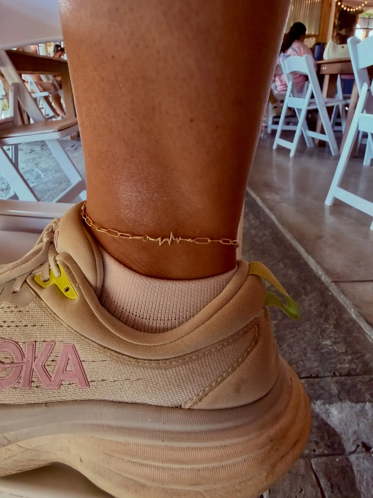 A gold chain anklet with a heartbeat charm, worn over a beige sneaker on a patio