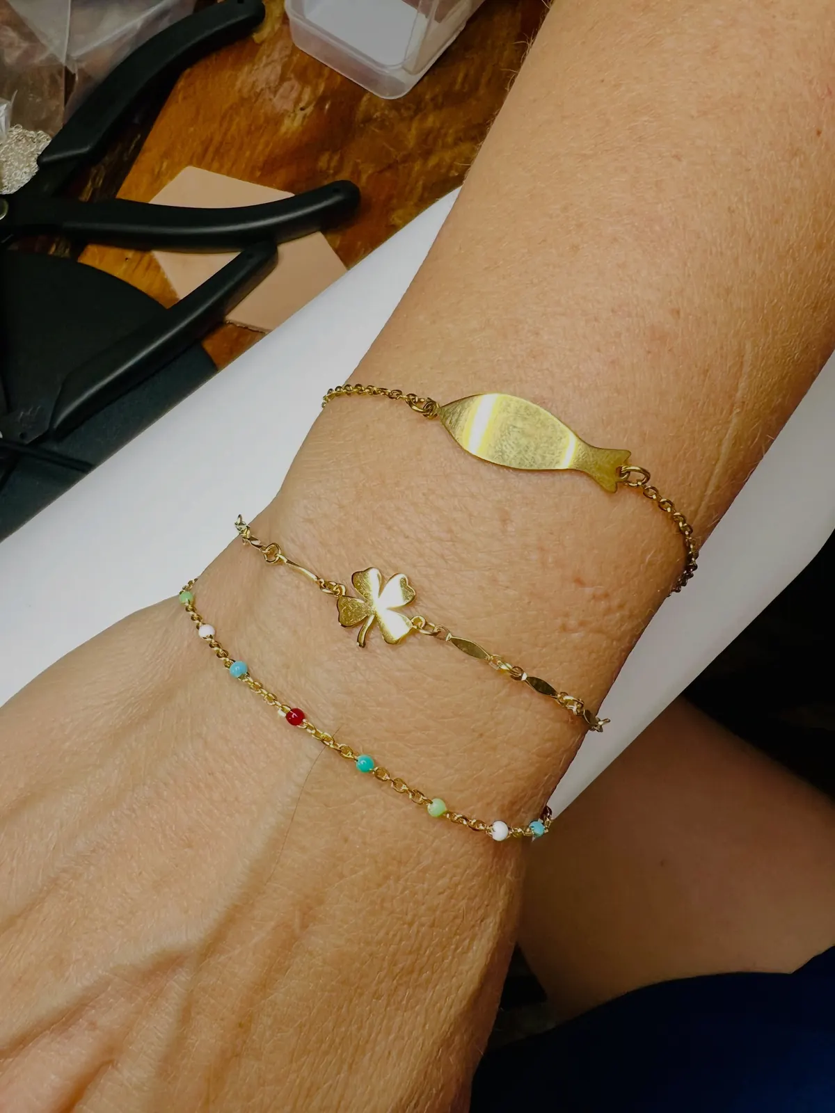 Three bracelets stacked on a forearm: a fish-shaped plate chain, a four-leaf clover chain and a multicolor bead chain