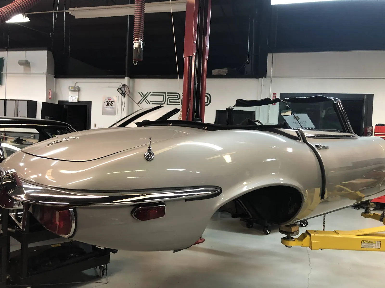 A silver Jaguar E-Type roadster body raised on a two-post lift in the GC Performance Classics shop, restoration work in progress