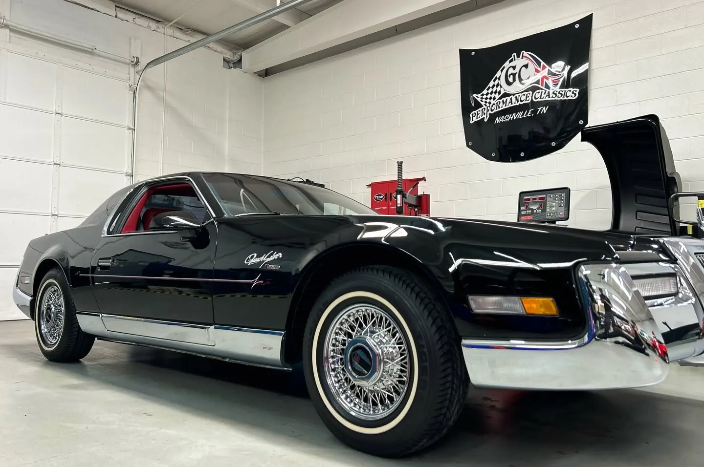 A black classic coupe with side pipes, wire wheels and whitewall tires, hood raised, in the shop under a GC Performance Classics banner