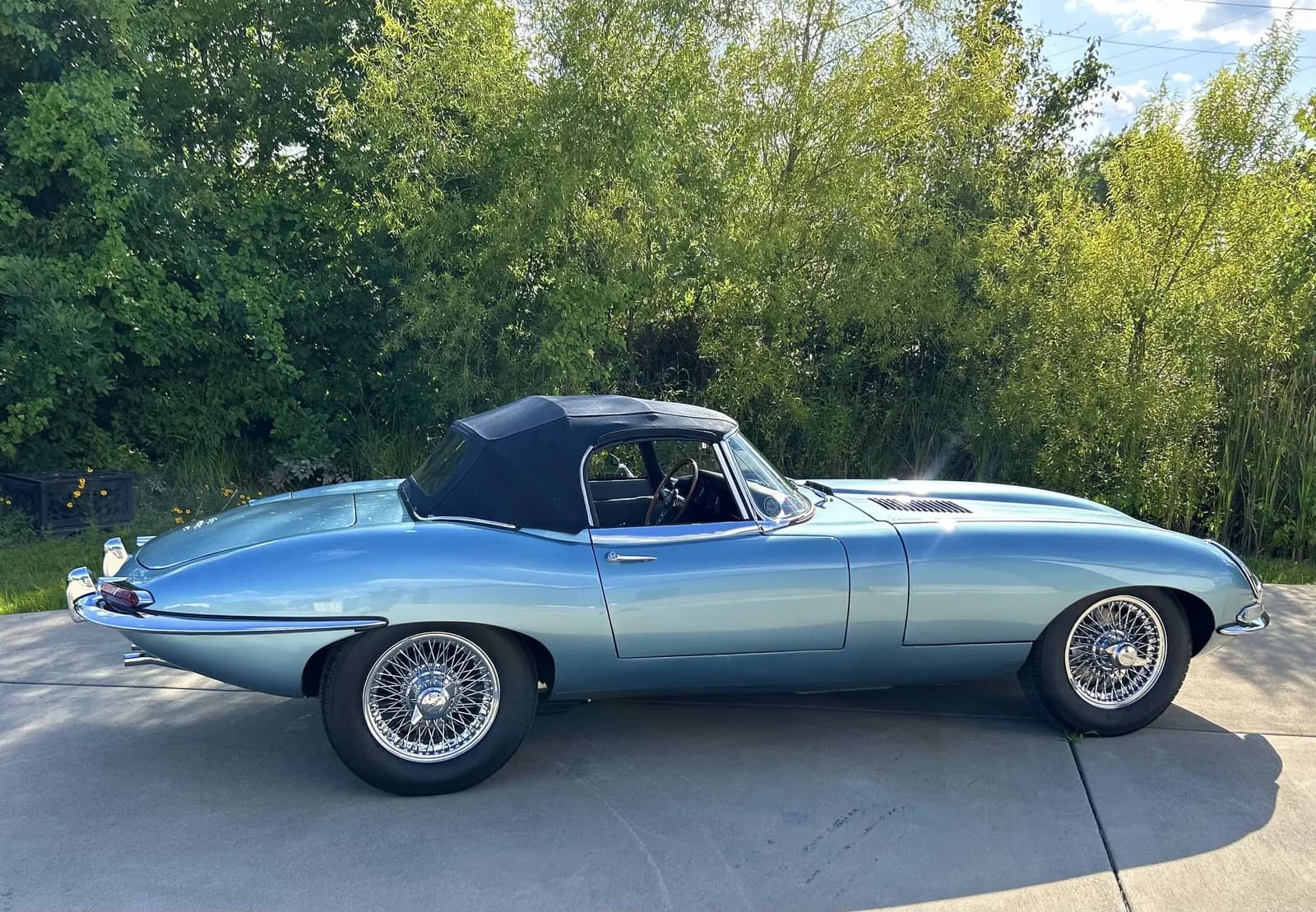 A light blue Jaguar E-Type roadster with its soft top up, in profile on a concrete apron with trees behind