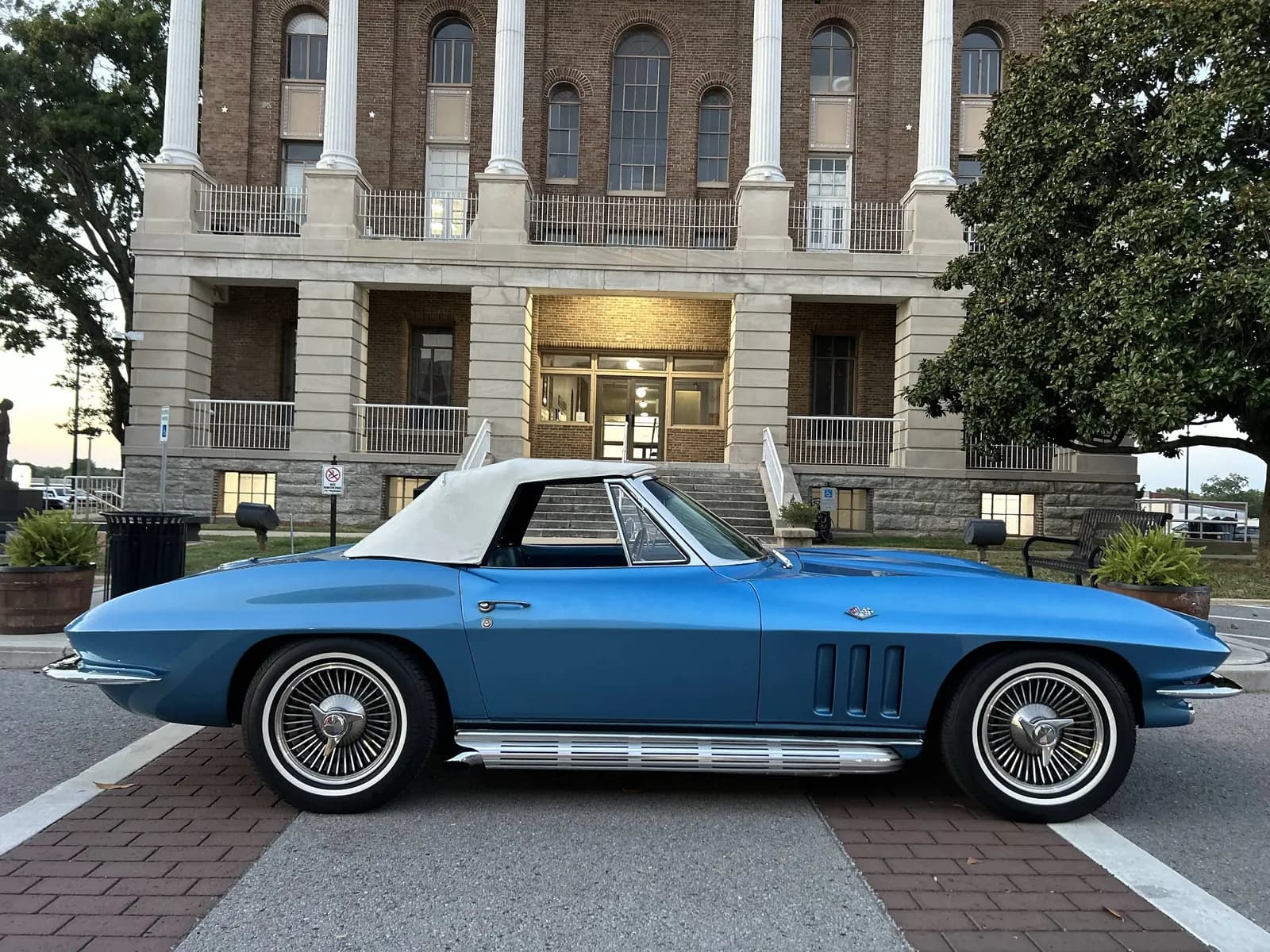 A blue 1960s Corvette Sting Ray convertible with a white top, parked in front of a brick courthouse