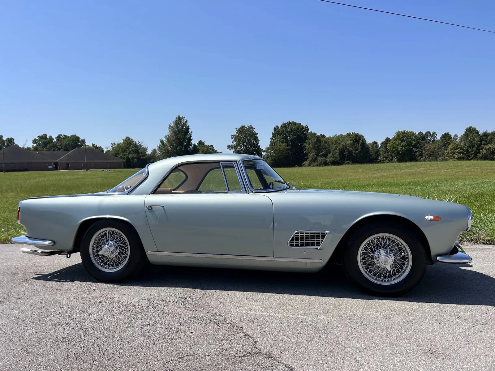 A silver blue Maserati coupe in side profile on a lot with a field and trees behind