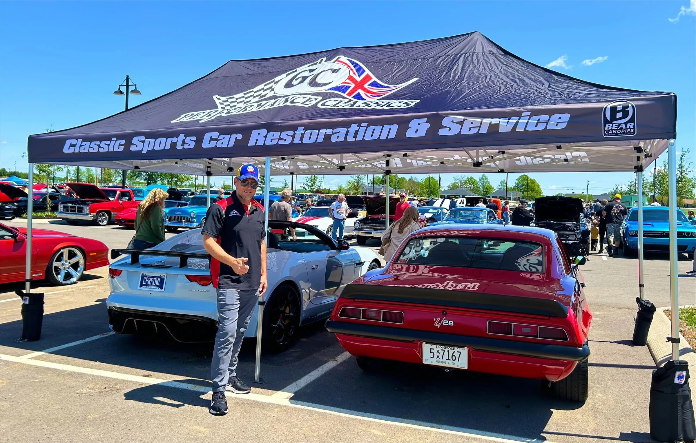 A GC team member gives a thumbs up under the GC Performance Classics canopy at a car show, between a white Jaguar F-Type and a red Camaro Z/28