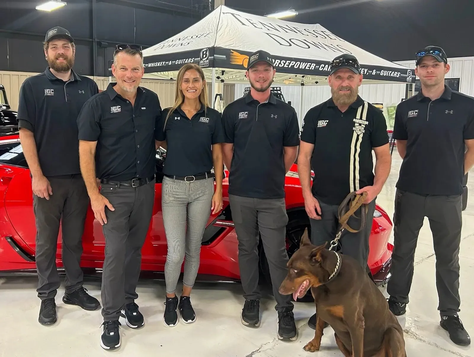 Six members of the GC Performance team in matching black polos, with a Doberman, in front of a red Corvette