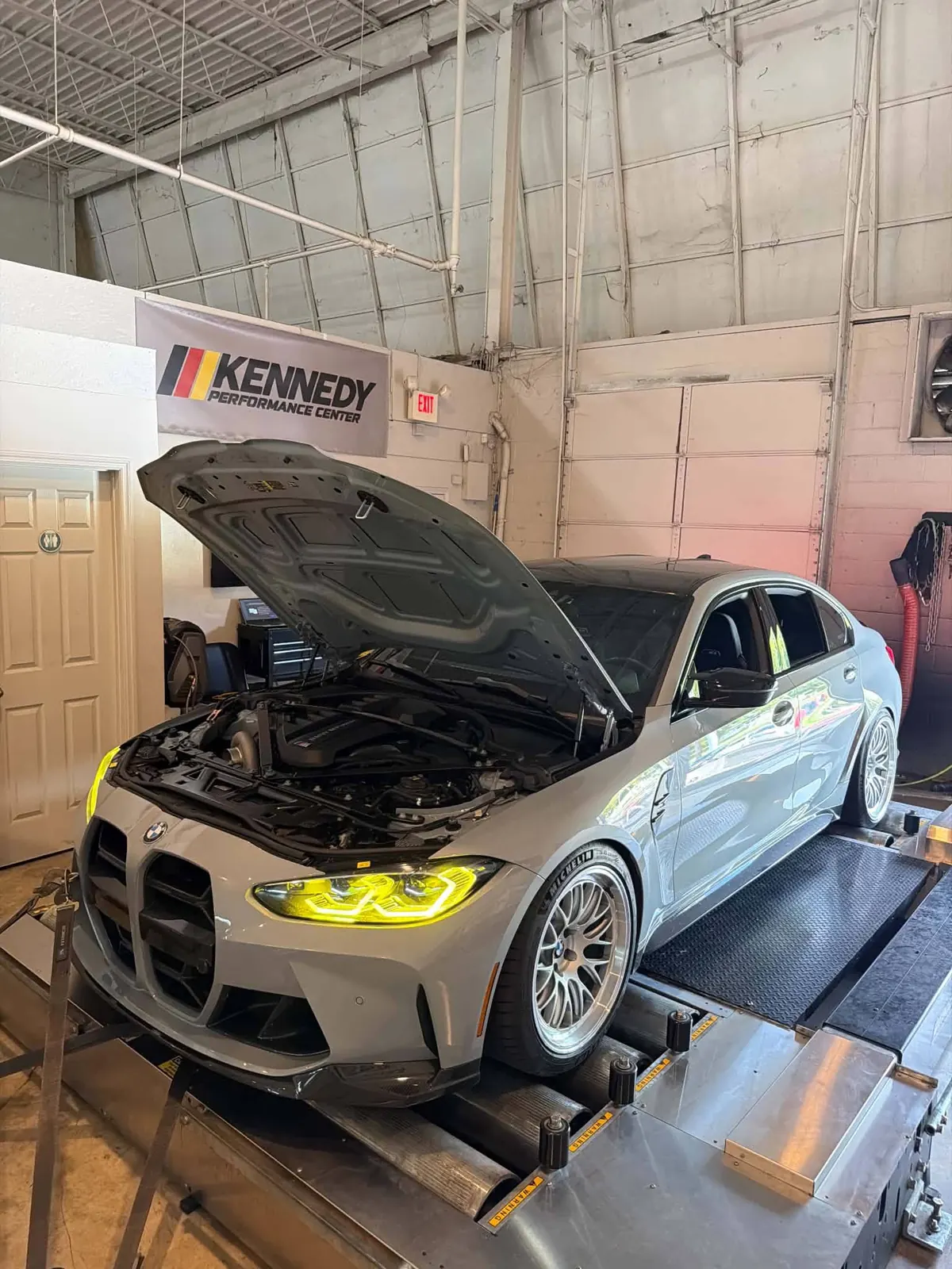 A gray BMW M3 sedan with its hood open on the dyno inside the Kennedy Performance Center shop, a Kennedy banner on the wall behind it