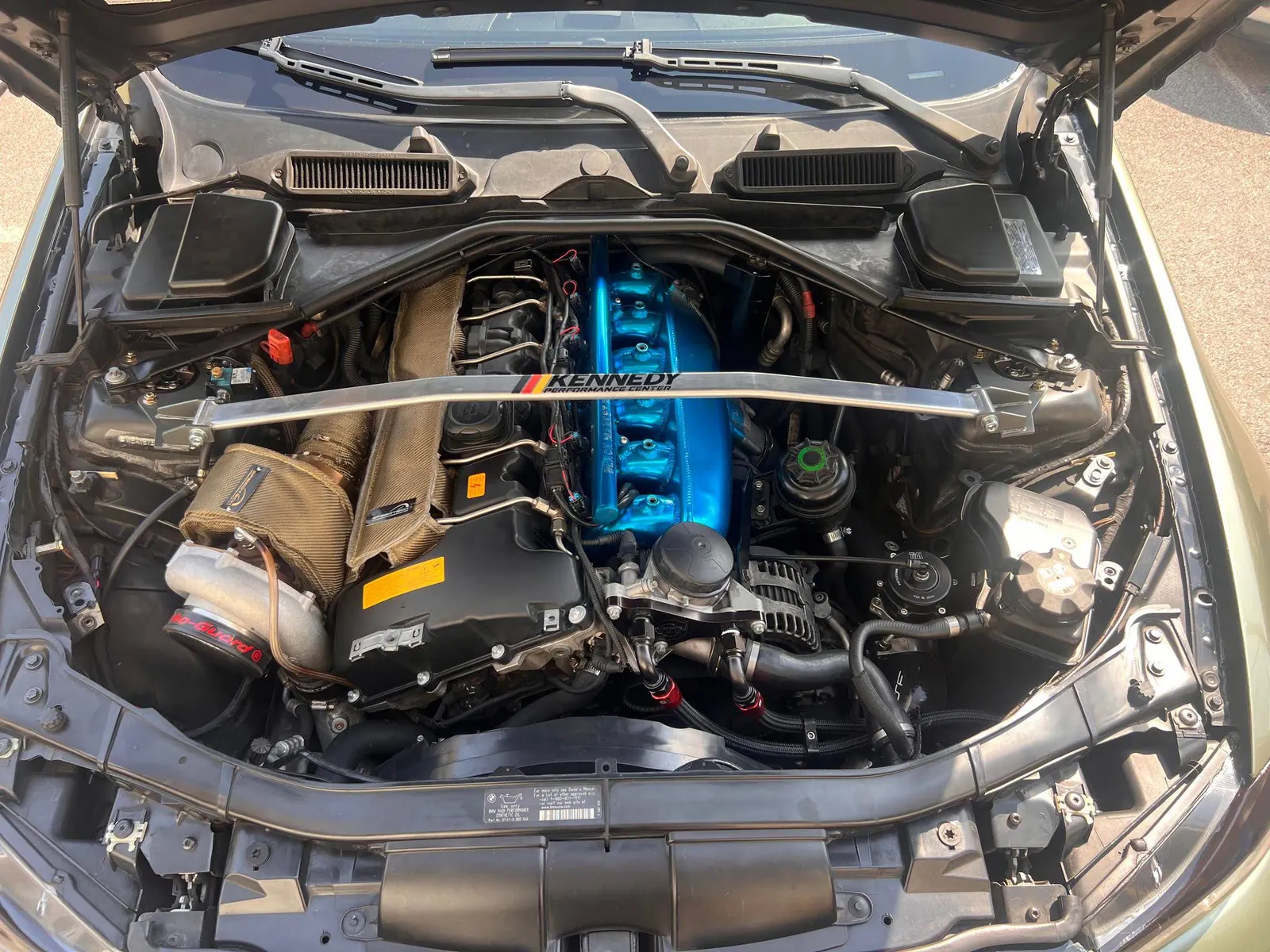 The engine bay of a BMW inline six with a blue painted top end, a gold wrapped pipe and a strut brace carrying the Kennedy logo