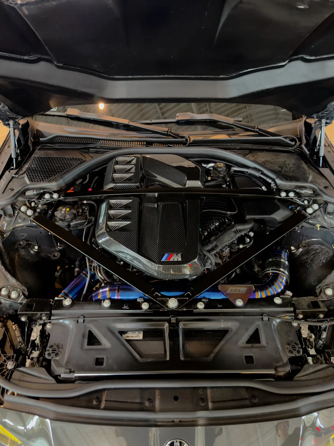 The engine bay of a current BMW M car with a carbon engine cover, an M badge and blue and gold titanium piping