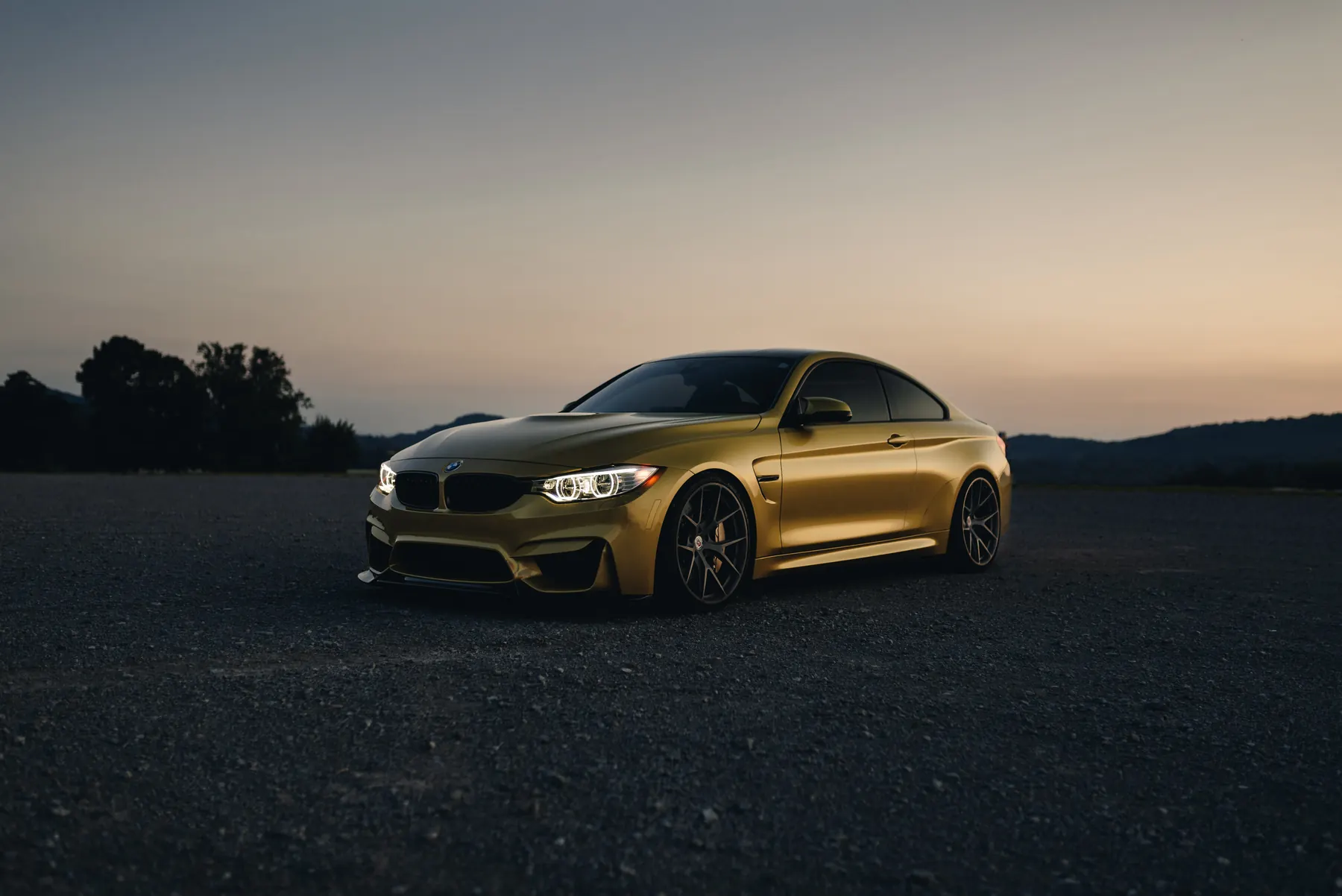 A gold BMW M4 coupe at dusk on an open lot, hills behind it