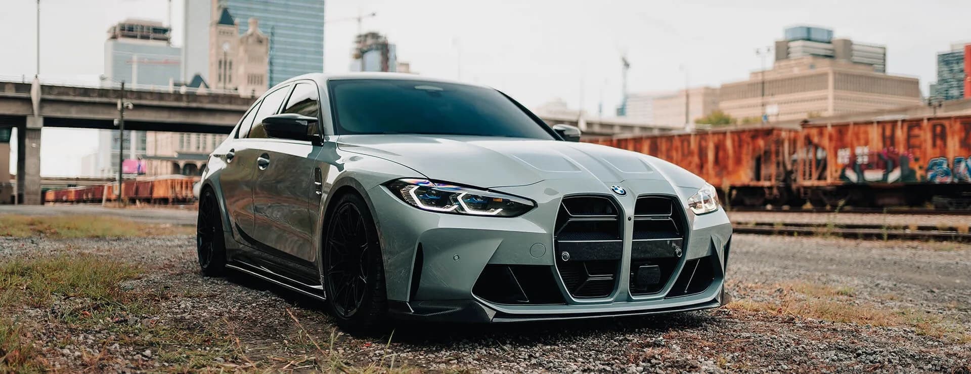 A gray BMW M3 sedan on gravel beside a rail yard, the city skyline behind