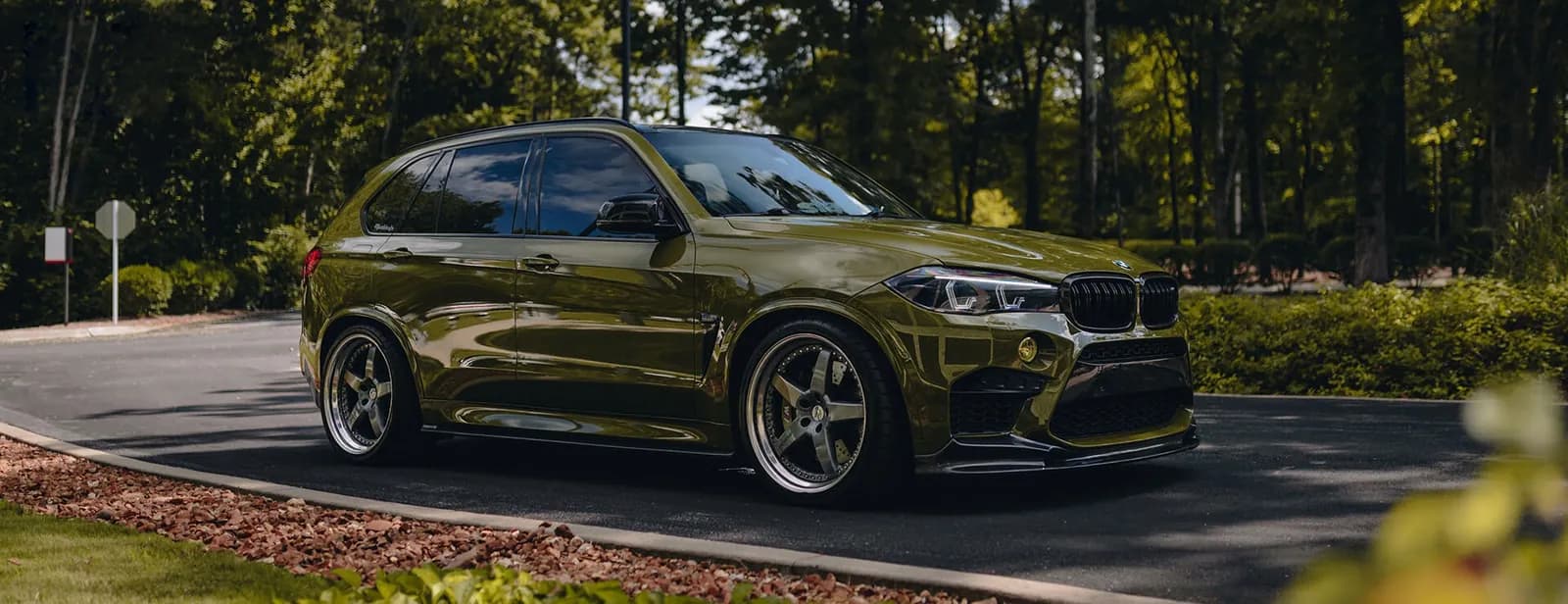 An olive green BMW X5 M on aftermarket wheels on a curving drive