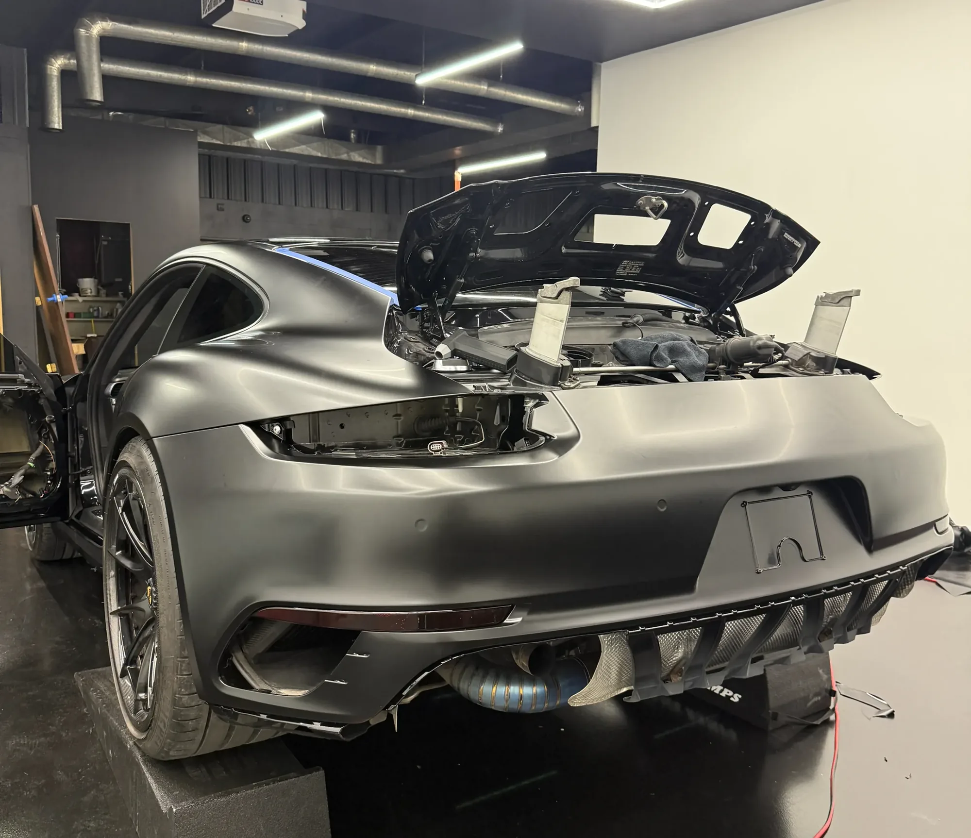 A Porsche 911 in satin black in the studio, its rear deck open mid-job, wiring and tools around it