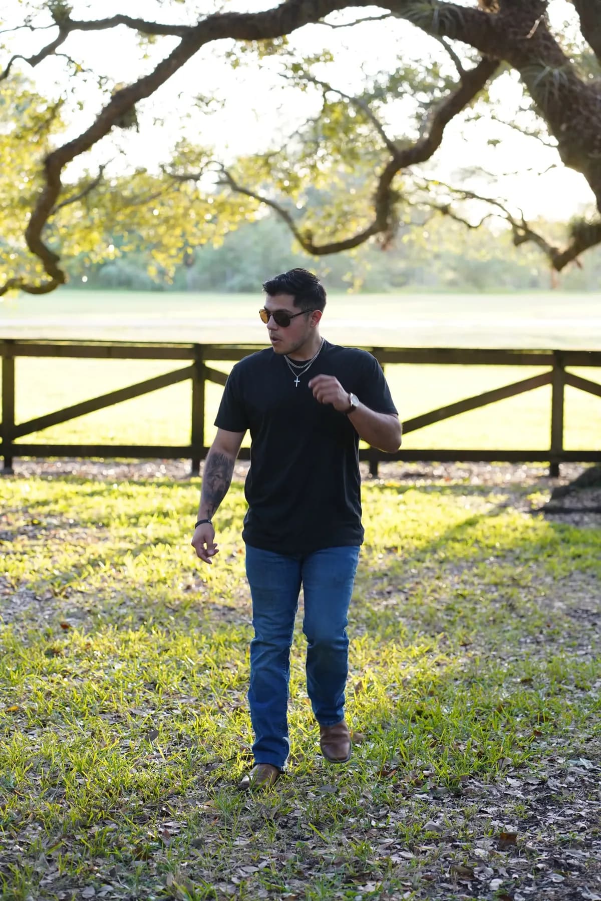 Andres Cueva, owner of La Cueva Auto Studio, walking across the grass by a wooden fence under a tree