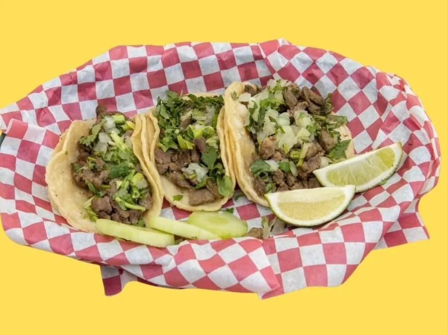 Three steak tacos on double corn tortillas with onion and cilantro, in a red and white checkered basket with lime wedges