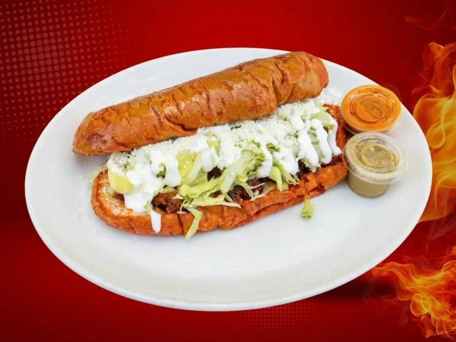 A pambazo, a red sauced sandwich on a long roll topped with lettuce and cream, on a white plate with two salsa cups