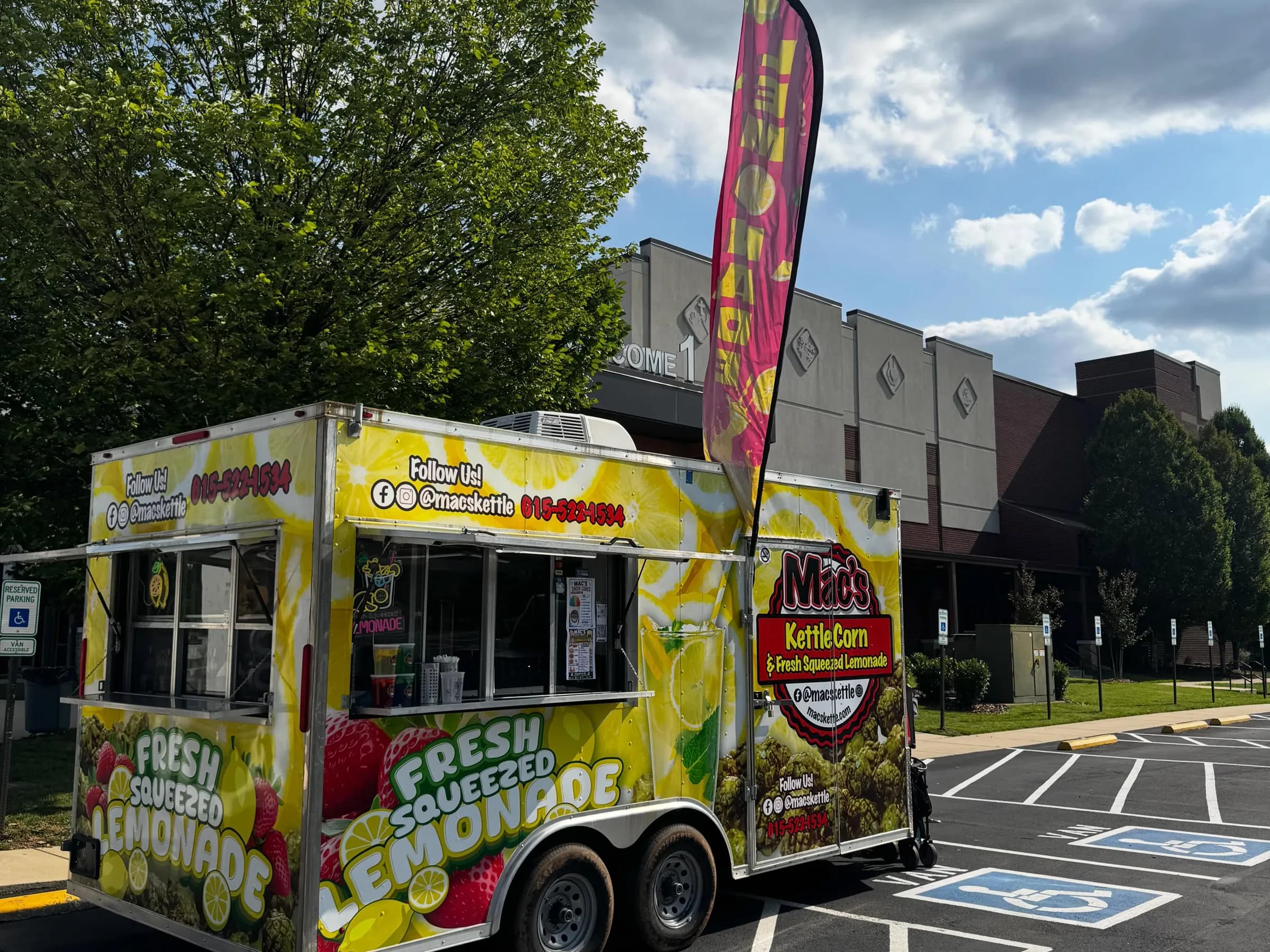 The Mac's trailer parked in daylight, wrapped in lemons and strawberries with the red Mac's emblem on its side, a Lemonade feather flag flying above it