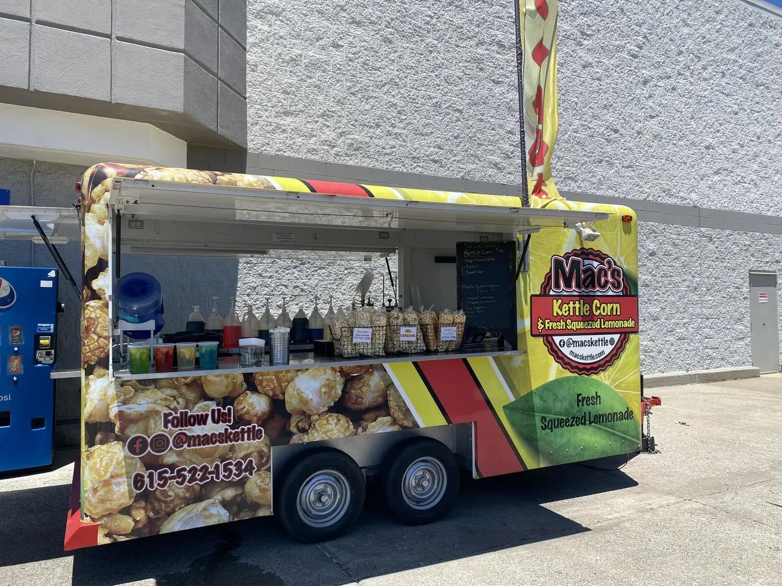 The Mac's trailer in the sun beside a store wall, its service window open over bagged kettle corn in wire baskets with flavor cards, a feather flag above it