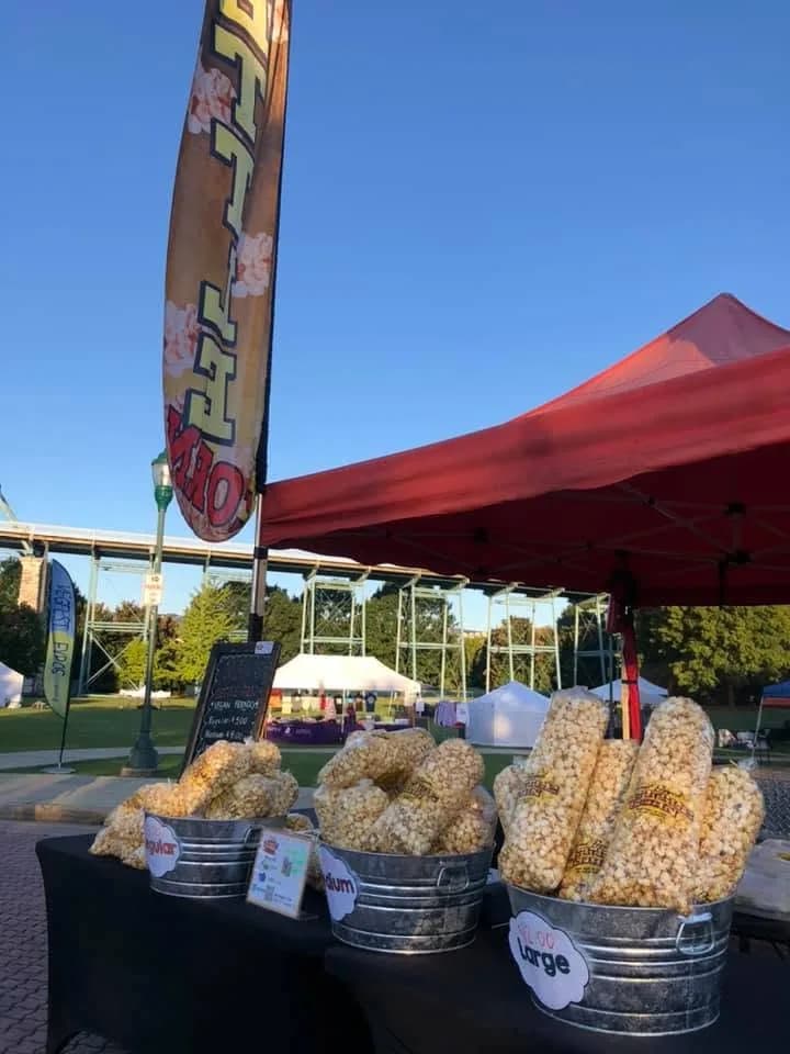 Three galvanized buckets of bagged kettle corn on a black table, tagged Regular, Medium and Large, under a red canopy