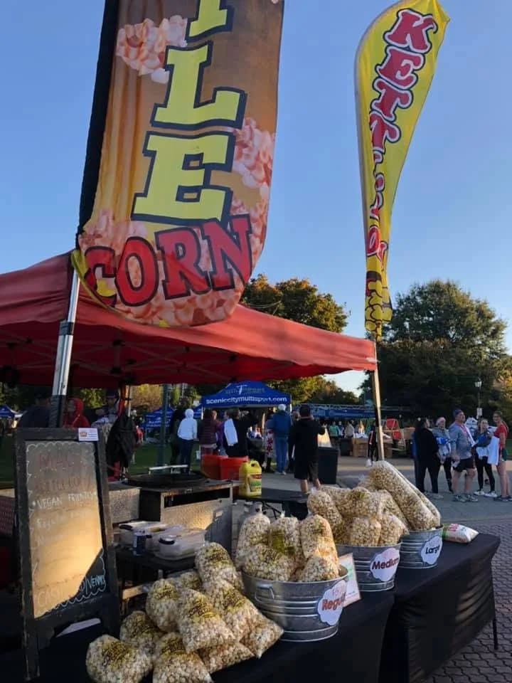 Bags of kettle corn piled in and around buckets on a black table, a chalkboard sign on an easel beside them, a red canopy and a Kettle Corn flag above