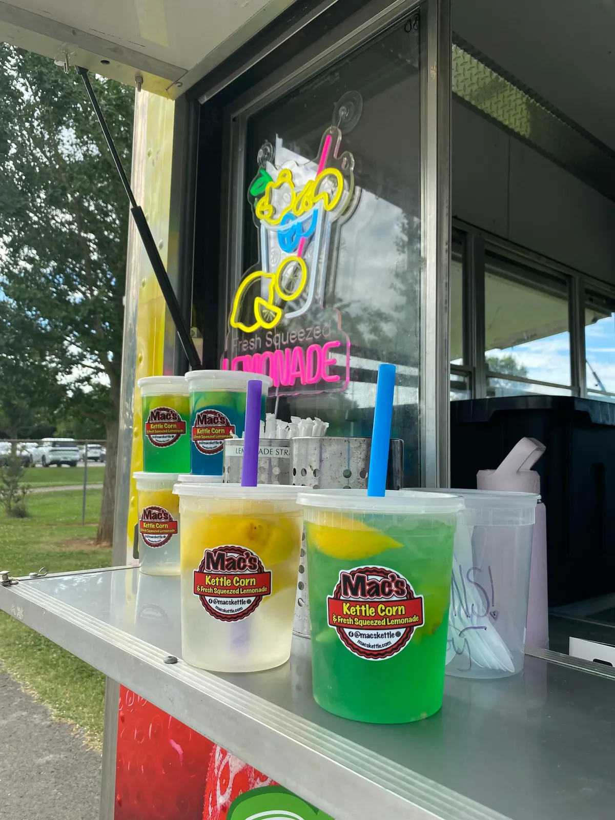 Lemonades in clear cups, yellow, green and blue, each with the Mac's sticker, on the stainless serving ledge beside the neon Fresh Squeezed Lemonade sign
