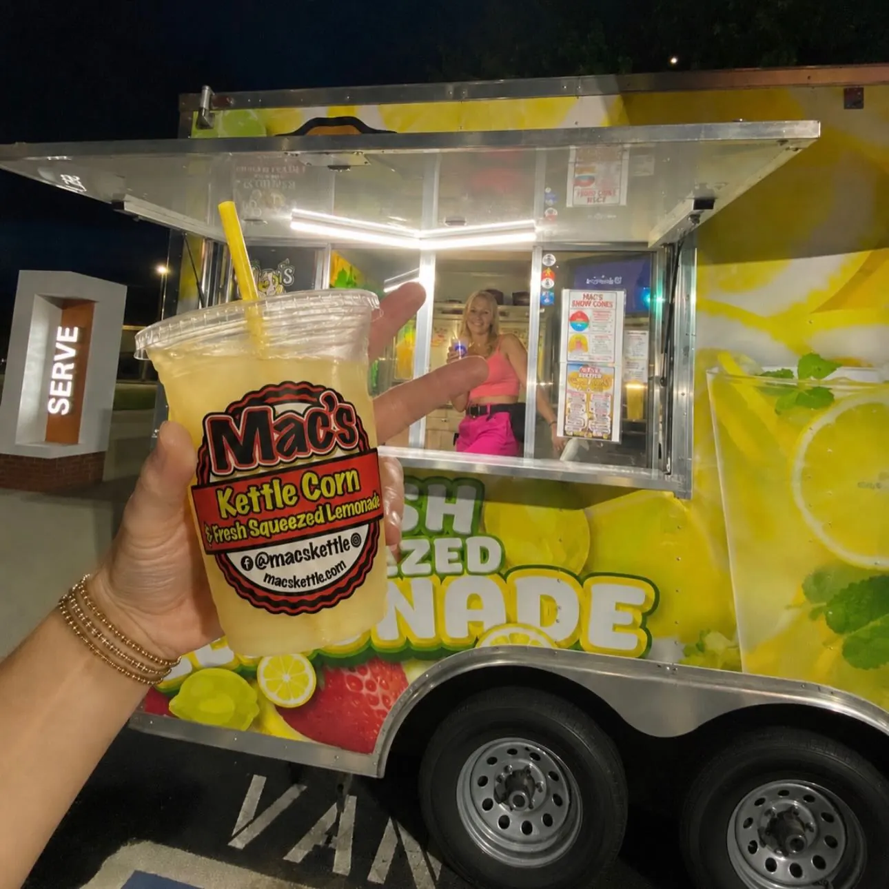 A hand holds a clear lemonade cup with the round Mac's sticker in front of the lit serving window