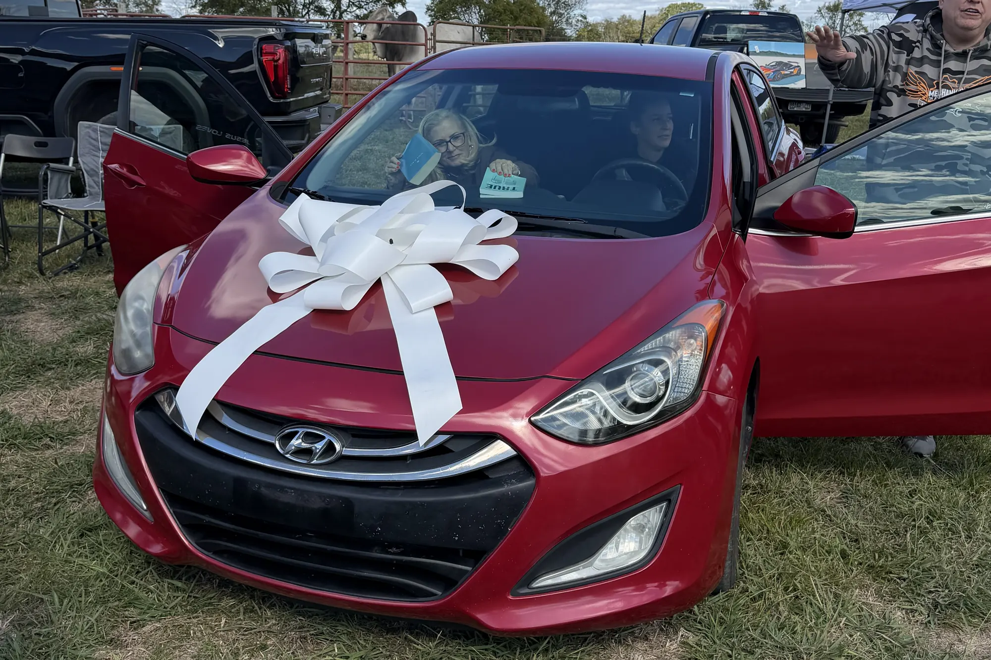 A red Hyundai hatchback with a big white bow on its hood, its doors open, on the grass at the ranch