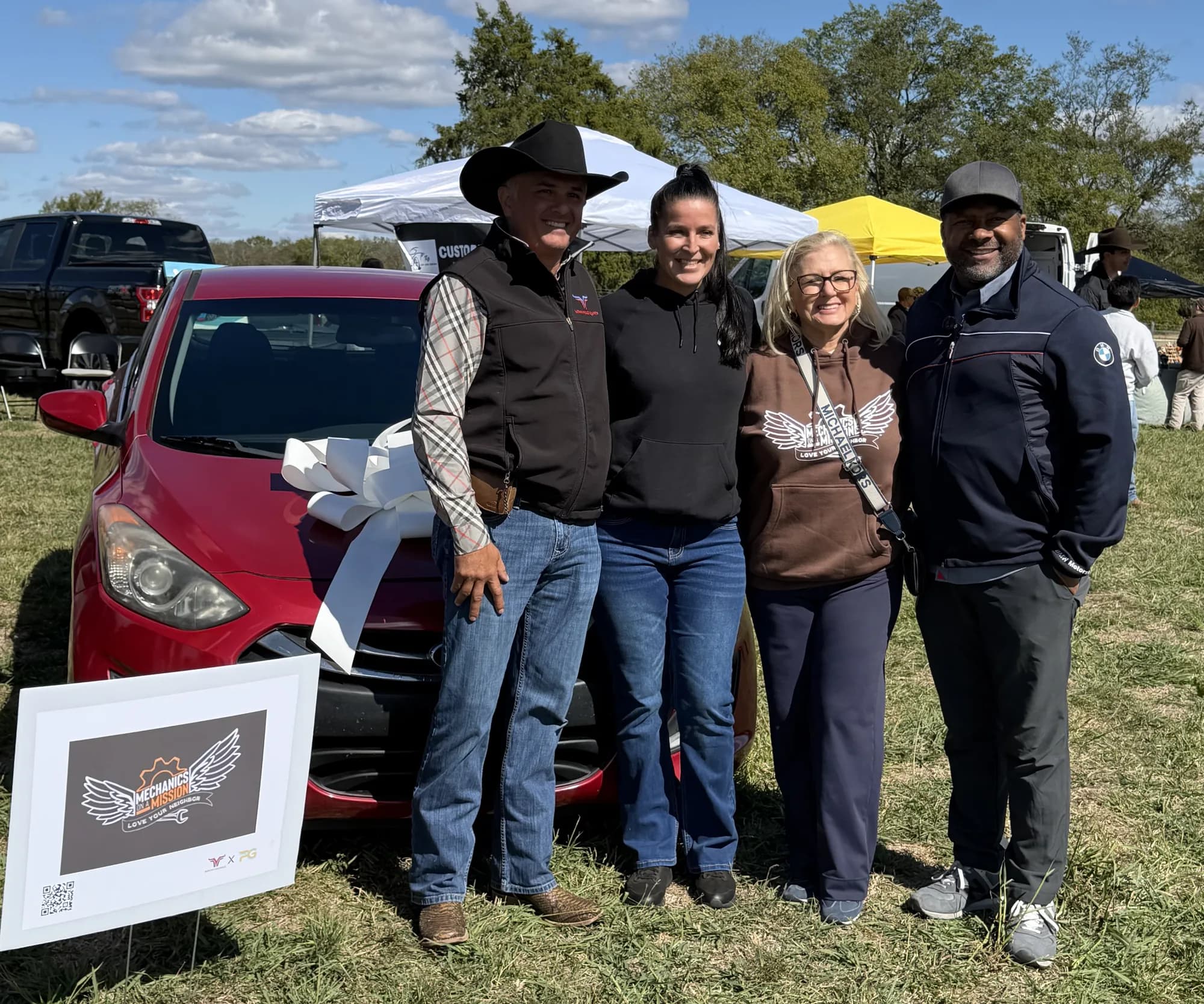 Four people standing together in front of the red car with the white bow, a framed Mechanics on a Mission print beside them