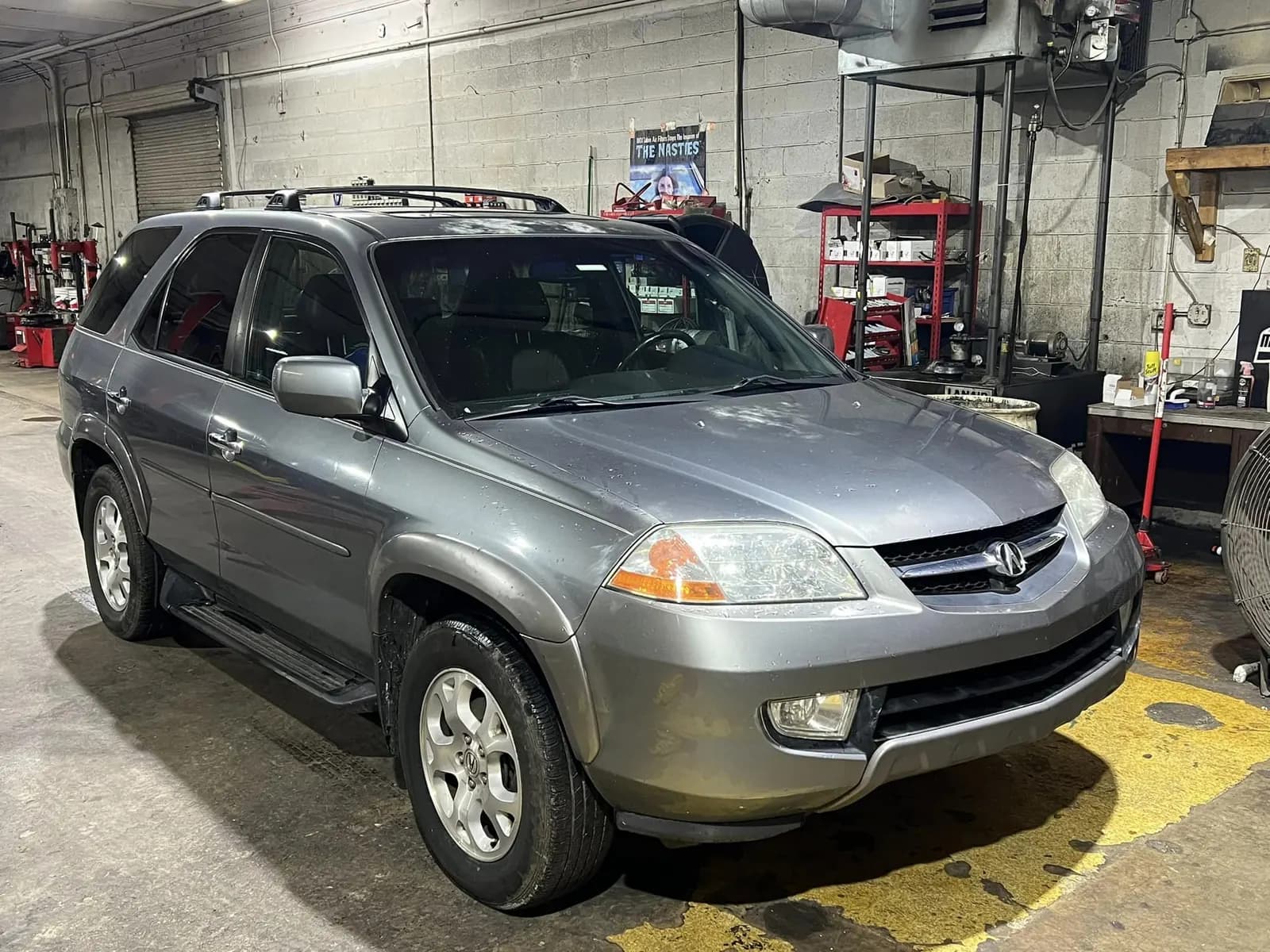 A gray Acura SUV parked in a repair shop bay, with tire machines, tool carts and parts shelves behind it
