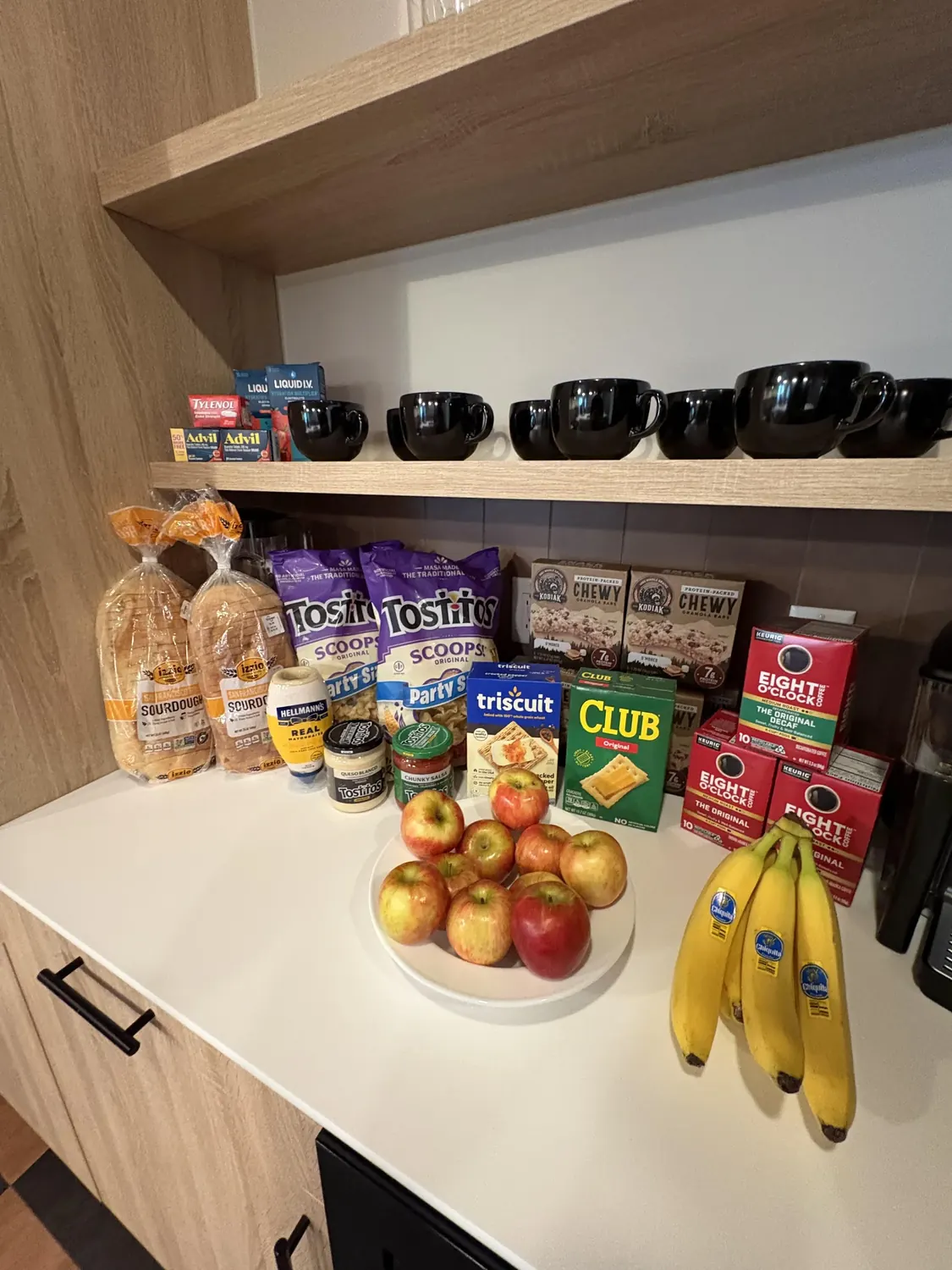 A white countertop with bread loaves, chips, crackers and snack boxes, a bowl of apples and a bunch of bananas