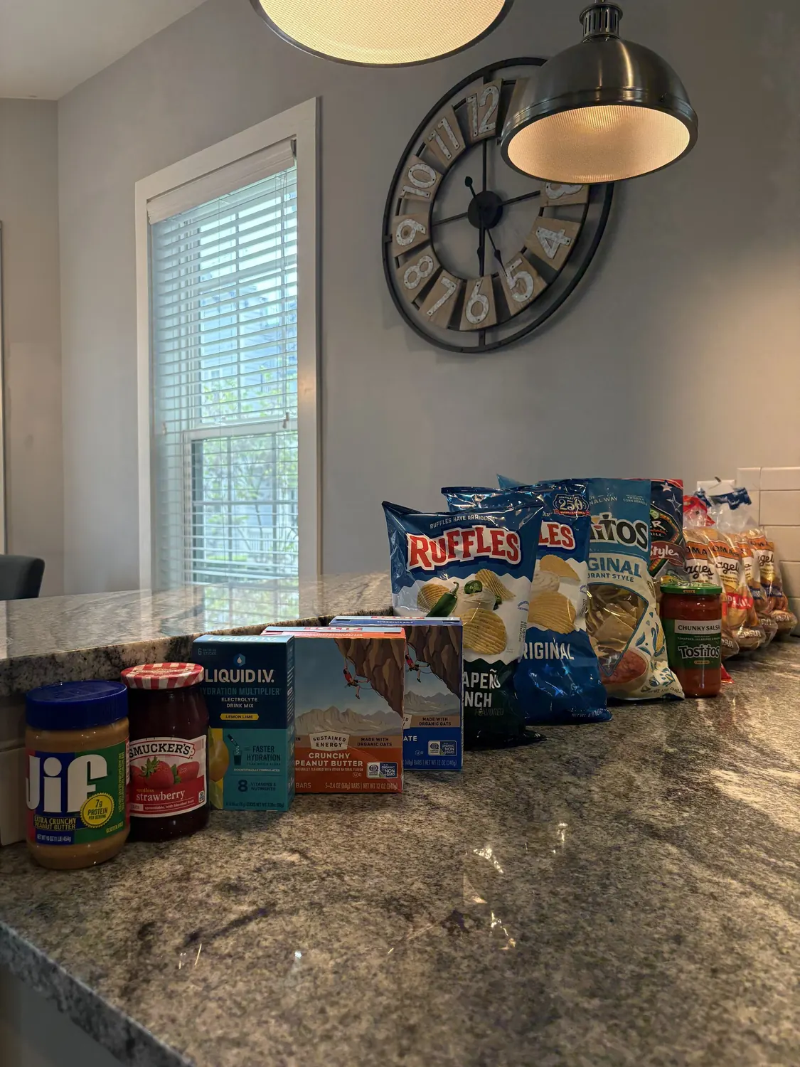 A granite counter with bags of chips, peanut butter, strawberry jam, electrolyte packets and bread