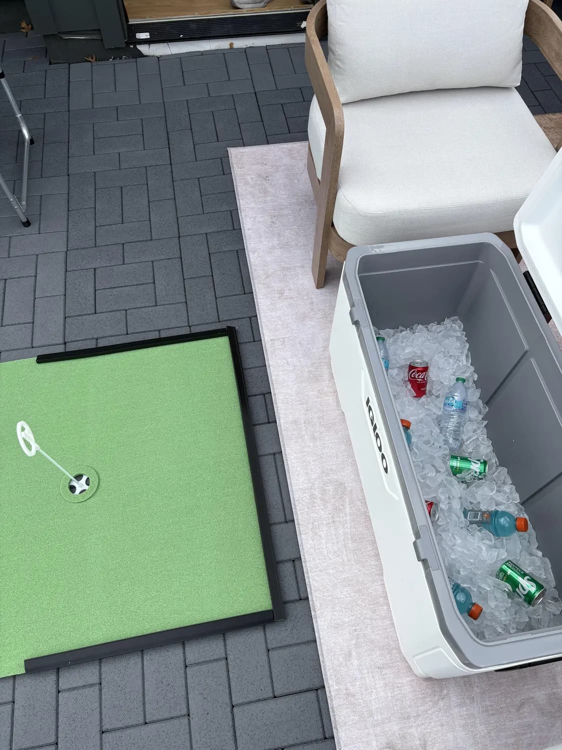 An Igloo cooler full of ice with soda, sports drinks and water, beside a roll out putting green on a paver patio