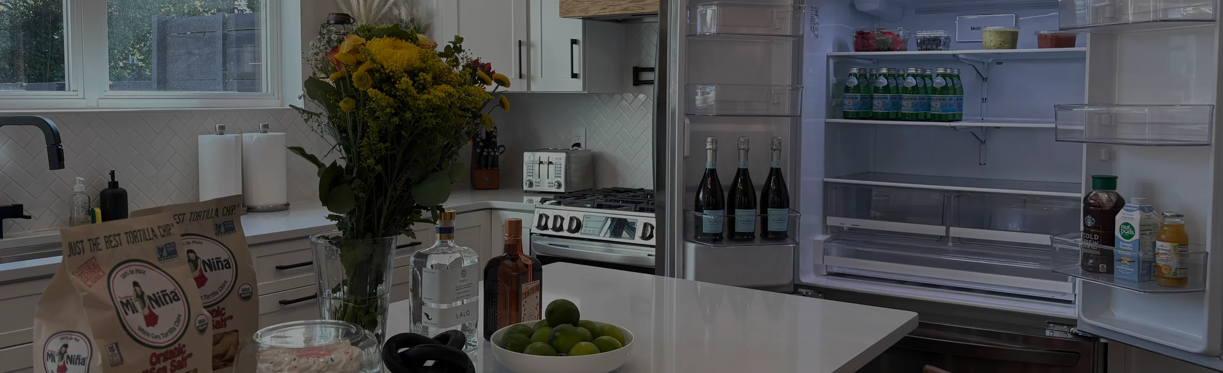 A kitchen with an open, stocked refrigerator, flowers, chips, limes and bottles on the island