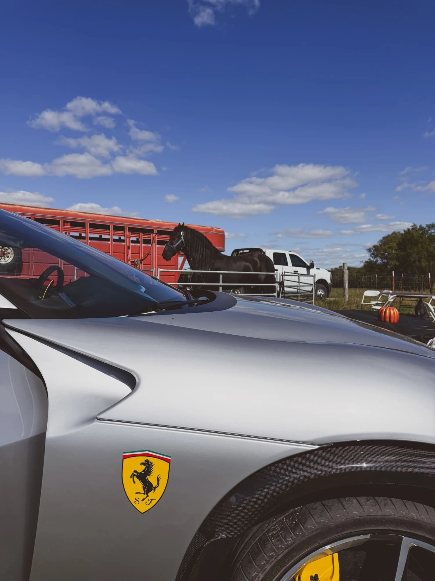 The Ferrari shield on the silver Purosangue's fender, and beyond it the ranch's black horse standing at the red livestock trailer