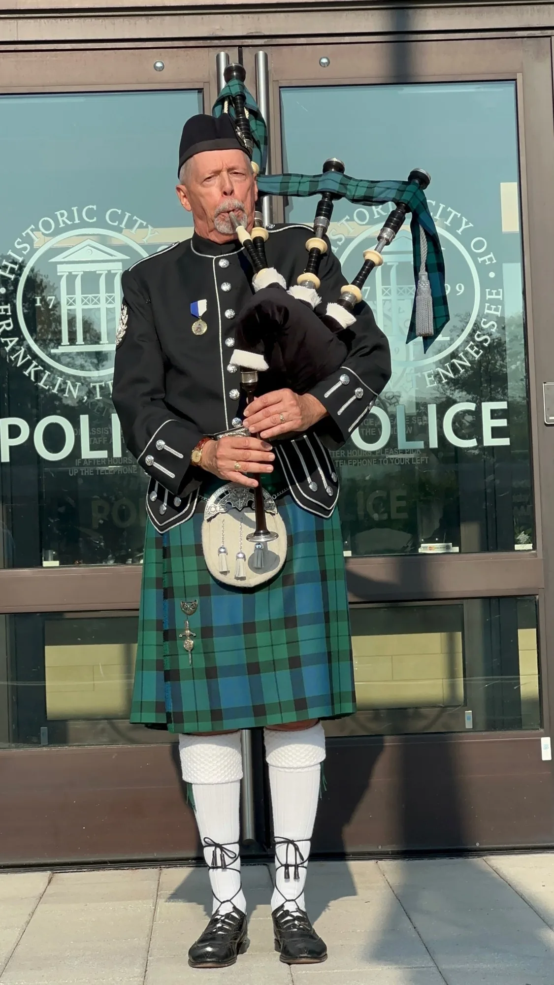 Richard Katz in Highland dress plays the bagpipes outside the glass doors of Franklin Police headquarters, the City of Franklin seal on the glass
