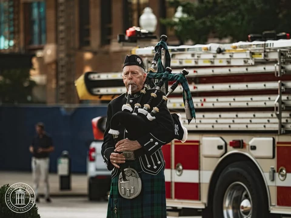 Richard Katz plays the bagpipes on a street, a red and white fire engine behind him