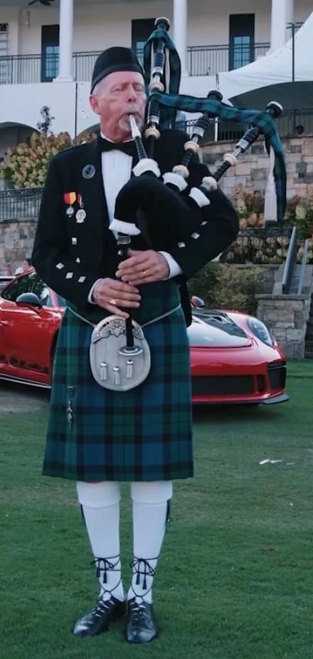 Richard Katz in Highland dress plays the bagpipes on a lawn, a red Porsche parked behind him