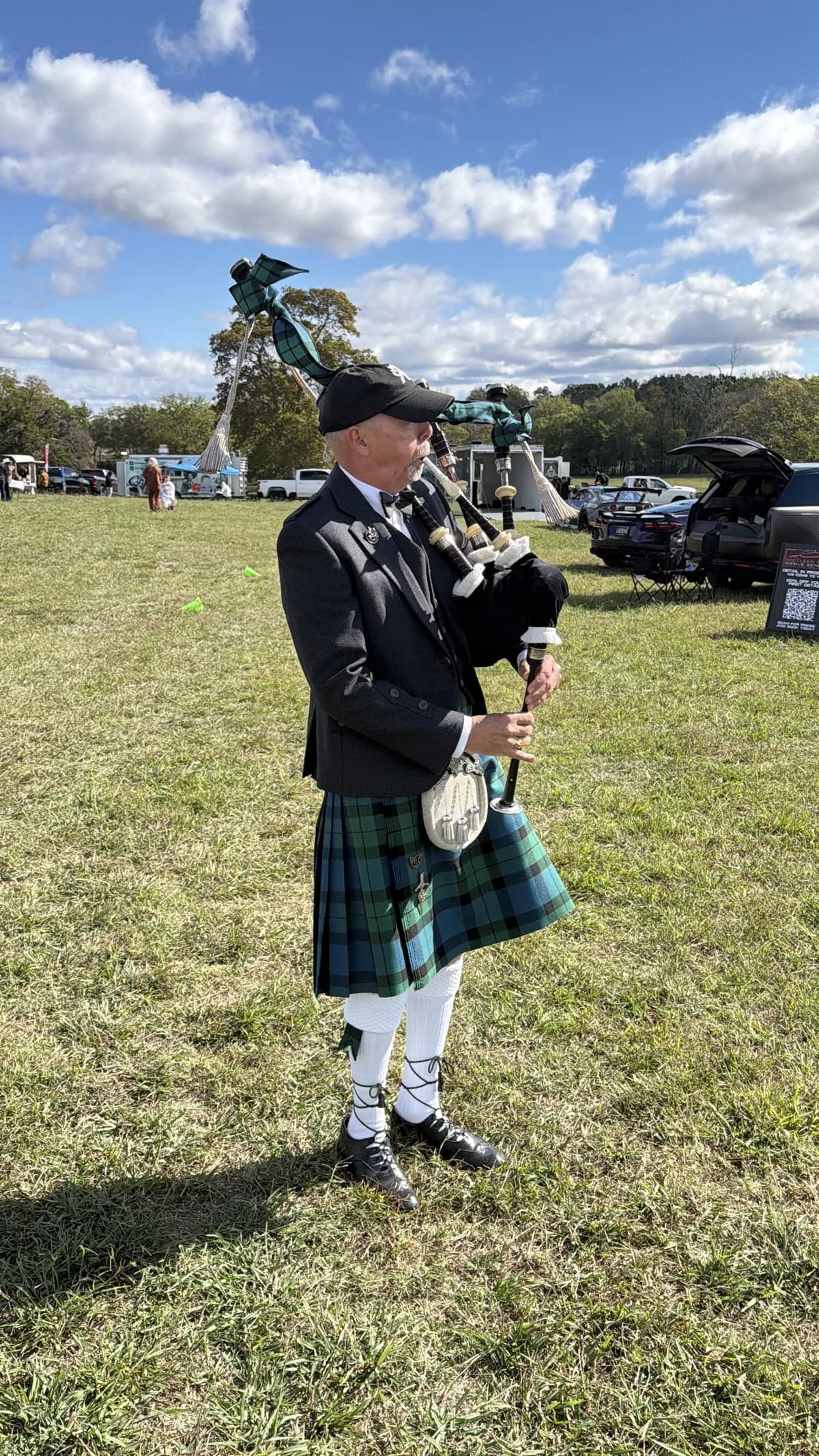 Richard Katz in Highland dress and a black cap plays the bagpipes on the show field, show cars parked behind him