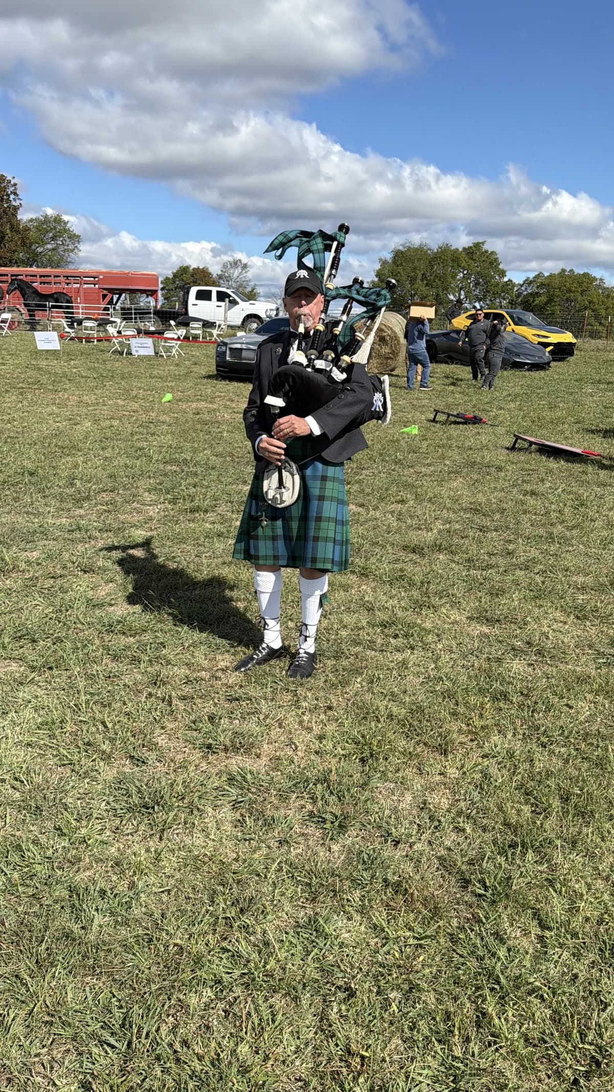 Richard Katz plays the bagpipes on the grass, a red livestock trailer and show cars behind him