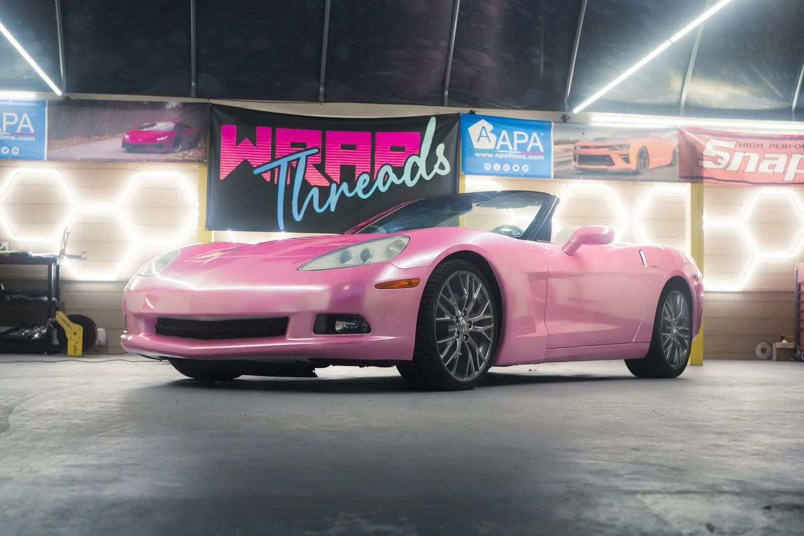 A pink Corvette convertible inside the Wrap Threads studio, under their neon logo banner and hexagon lights