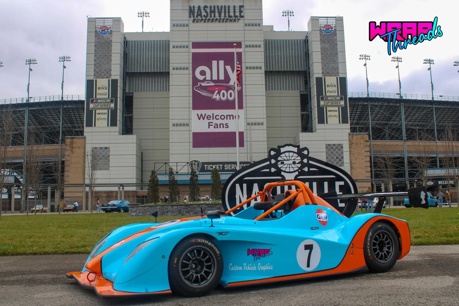 A turquoise and orange race car, number 7, in front of the grandstand at Nashville Superspeedway