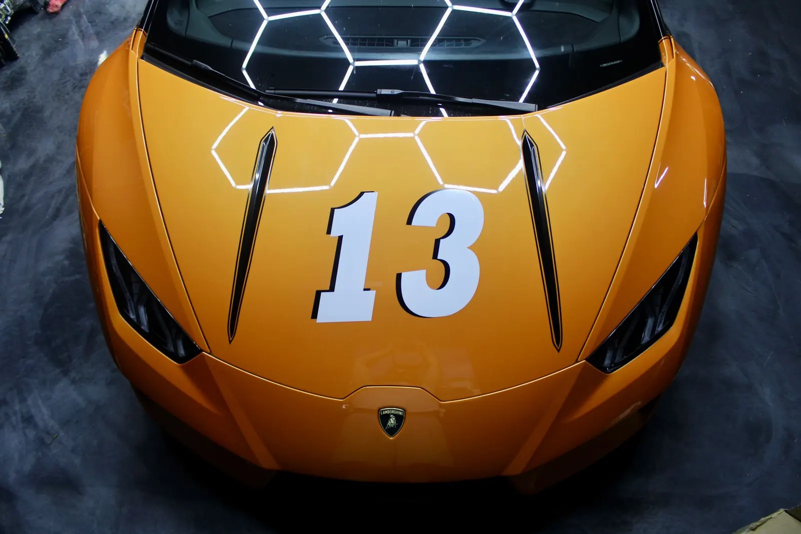 The hood of an orange Lamborghini with a white number 13, the shop's hexagon lights reflected in it